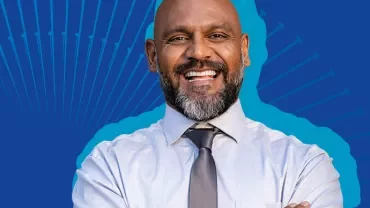 Homem sorrindo em fundo azul, vestindo camisa social.